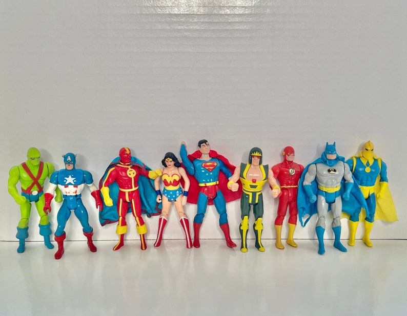 May include: A collection of eight superhero action figures, including Superman, Batman, Wonder Woman, The Flash, Captain America, Green Lantern, and two others. The figures are all in various poses and are made of plastic.