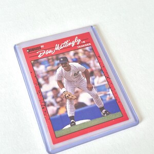 May include: A baseball card featuring Don Mattingly of the New York Yankees. The card has a red border and the text "Don Mattingly" and "Yankees" is visible. The card is encased in a clear plastic holder.