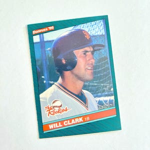 May include: A baseball card depicting Will Clark in a San Francisco Giants uniform. The card has a teal border with the text "Donruss '86" at the top and "The Rookies" and "WILL CLARK 1B" at the bottom. The image shows Clark wearing a batting helmet.