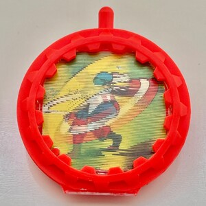 May include: Red plastic toy with a gear-shaped rim and a clear plastic window showing a cartoon image of a superhero in a red, white, and blue costume.