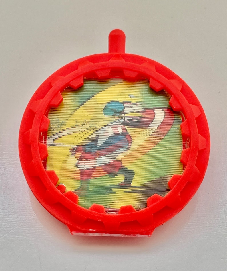 May include: Red plastic toy with a gear-shaped rim and a clear plastic window showing a cartoon image of a superhero in a red, white, and blue costume.