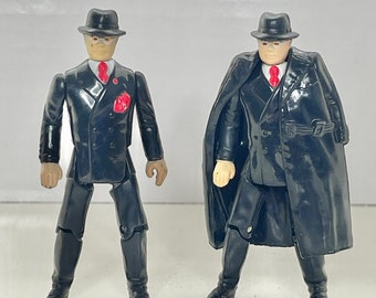 May include: Two black plastic action figures of men wearing black suits, white shirts, red ties, and black hats. One figure is wearing a black coat.