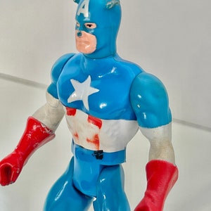 May include: A vintage action figure of Captain America in a blue and red costume with a white star on his chest. The figure has a red and white helmet with the letter "A" on the front.
