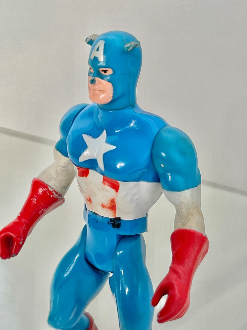 May include: A vintage action figure of Captain America in a blue and red costume with a white star on his chest. The figure has a red and white helmet with the letter "A" on the front.