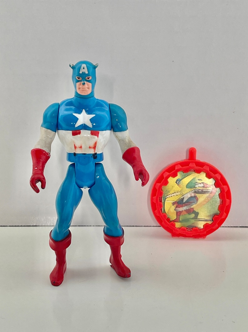 May include: A vintage action figure of Captain America in his classic blue, red, and white costume. The figure has a red, white, and blue shield with a star on it. The figure is holding a red and white shield. The figure is standing on a white background. The figure is in good condition.