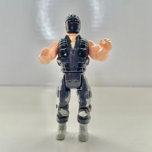 May include: A vintage action figure of a soldier in a black helmet and vest, with gray and white camouflage pants and boots. The figure has tan arms and is posed with arms raised. The toy is standing on a white surface.