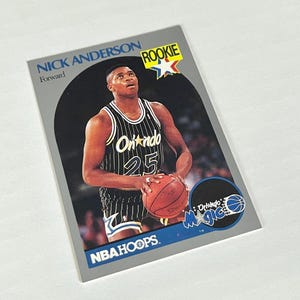 May include: A Nick Anderson rookie basketball card from NBA Hoops. The card features Anderson in an Orlando Magic uniform, holding a basketball. The card has a gray border and the text "ROOKIE" in the upper right corner.