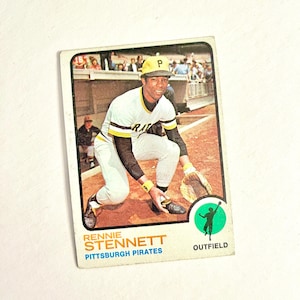 May include: A vintage baseball card featuring Rennie Stennett of the Pittsburgh Pirates. The card shows Stennett in his baseball uniform, with the team's logo and the word "OUTFIELD" visible. The card has a white border and a slightly worn appearance.