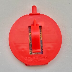 May include: Red plastic toy with a pull-out handle and a picture of a cartoon character. The toy has the text "MATTEL INC. 1983" and "HONG KONG" printed on the bottom.