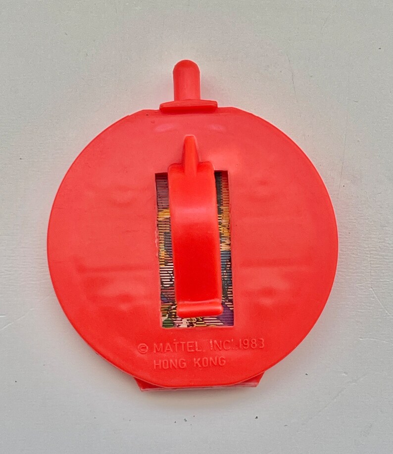 May include: Red plastic toy with a pull-out handle and a picture of a cartoon character. The toy has the text "MATTEL INC. 1983" and "HONG KONG" printed on the bottom.