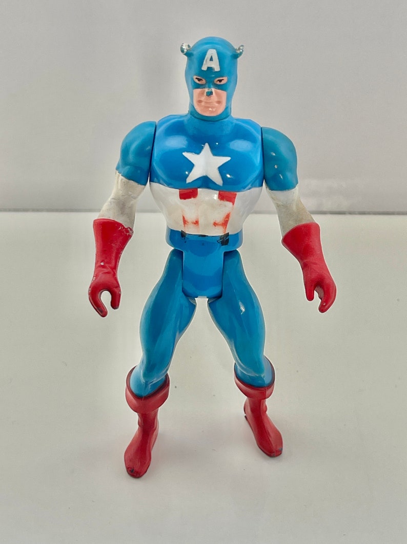 May include: A vintage action figure of Captain America, wearing his iconic red, white, and blue costume. The figure has a blue body, red gloves, and red boots. The figure has a white star on its chest and the letter 'A' on its forehead.