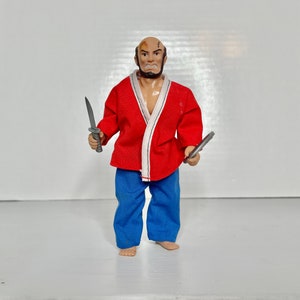 May include: A vintage action figure of a bald, bearded man in a red jacket and blue pants, holding two silver knives. The figure has a stern expression and visible scars. The jacket has white trim.