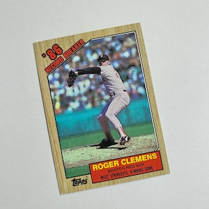 May include: A vintage baseball card featuring Roger Clemens of the Boston Red Sox. The card has a wooden-like border with the text "'86 RECORD BREAKER." The card also includes the Topps logo and details about Clemens' achievements.
