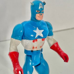 May include: A vintage action figure of Captain America, wearing a blue and red costume with a white star on his chest. The figure has a blue helmet with a white "A" on the front.
