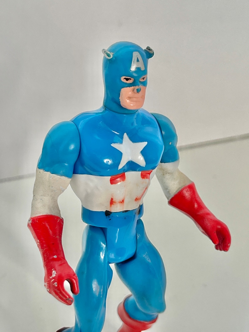May include: A vintage action figure of Captain America, wearing a blue and red costume with a white star on his chest. The figure has a blue helmet with a white "A" on the front.