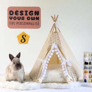 Pode incluir: Um pequeno tipi de tecido bege com padrão geométrico dourado, acabamento de pompons brancos e postes de madeira. Um coelho cinzento está sentado ao lado num tapete de pele sintética branca. Um sinal diz DESIGN YOUR OWN TIPI PERSONNALISÉ.