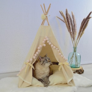 Puede incluir: Un tipi beige para gatos con un cojín blanco en el interior. El tipi tiene un ribete de pompones rosas y lazos amarillos. Un gato está sentado dentro del tipi.