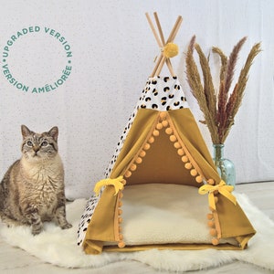 May include: A yellow and white patterned teepee-style pet bed with a white faux fur cushion. The teepee has yellow pom-poms around the bottom edge and yellow ribbon ties. A cat is sitting in front of the bed.