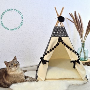 May include: A black and white geometric patterned teepee pet bed with a cream colored interior and black pom pom trim. The teepee is on a white faux fur rug with a tabby cat lying on the rug in front of the teepee.