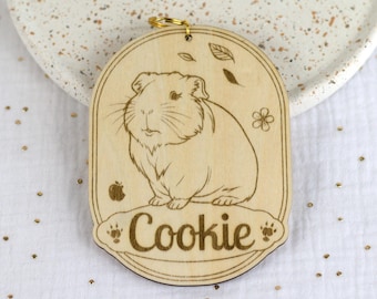 May include: A light-colored wooden oval ornament featuring a laser-engraved illustration of a guinea pig. The design includes small leaf and flower accents, a tiny apple icon, and the name Cookie written in a script font with paw print details.