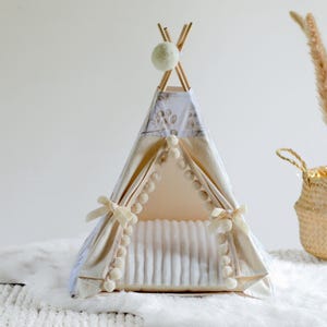May include: A small pet teepee tent featuring a cream-colored fabric with a subtle gray botanical print. The entrance is framed by white pom-pom trim and tied back with cream ribbons. A single white pom-pom hangs from the top wooden pole structure.