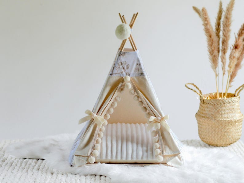May include: A small pet teepee tent featuring a cream-colored fabric with a subtle gray botanical print. The entrance is framed by white pom-pom trim and tied back with cream ribbons. A single white pom-pom hangs from the top wooden pole structure.