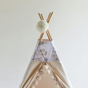 May include: A teepee tent featuring light beige fabric panels with a white pom-pom trim along the front opening. The top section displays a botanical print in muted brown tones, and the structure is topped with wooden poles and a white pom-pom.