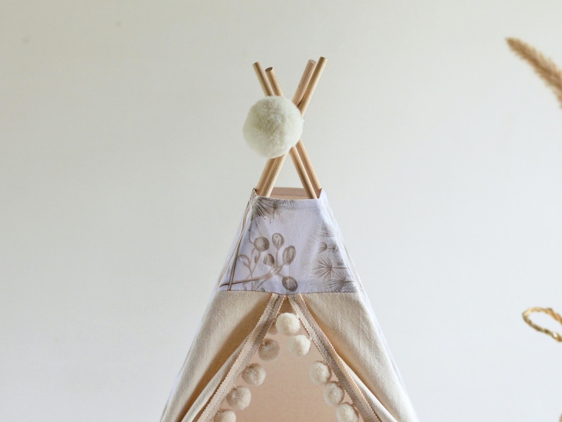 May include: A teepee tent featuring light beige fabric panels with a white pom-pom trim along the front opening. The top section displays a botanical print in muted brown tones, and the structure is topped with wooden poles and a white pom-pom.