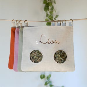 May include: A row of colorful hay bags hanging from a wooden rod. The bags are in shades of orange, pink, green, and white. The white bag has the name "Léon" in gold script and two circular openings filled with hay.