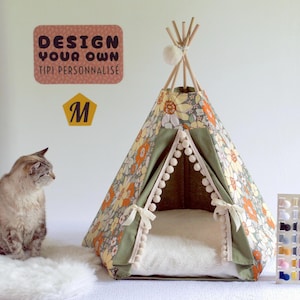 Puede incluir: Una cama para mascotas con forma de tipi con estampado floral y un cojín blanco en el interior. La tienda tiene postes de madera y está adornada con pompones blancos y tela verde. Un gato está sentado en una alfombra blanca junto a la cama. El texto "DESIGN YOUR OWN TIPI PERSONNALISÉ" es visible.