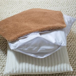 Puede incluir: Una funda de almohada de piel sintética marrón con cierre de cremallera se coloca sobre una almohada blanca con una funda de almohada blanca acanalada.