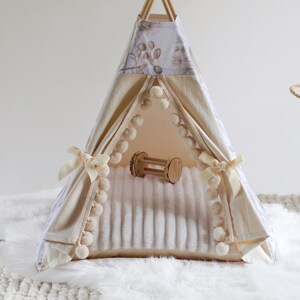 May include: A small teepee tent featuring cream-colored fabric with a subtle botanical print, accented by cream pom-pom trim along the front opening and tied with matching ribbons. Inside, a soft white textured mat holds a small wooden toy.