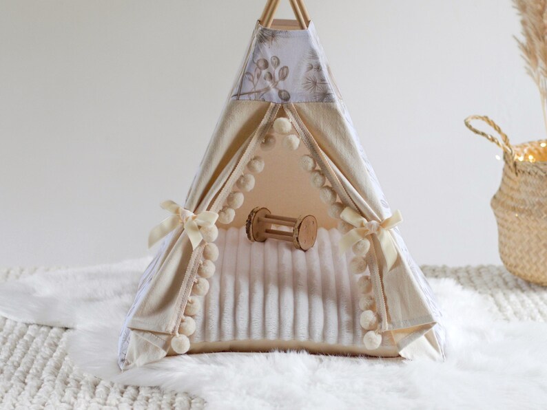May include: A small teepee tent featuring cream-colored fabric with a subtle botanical print, accented by cream pom-pom trim along the front opening and tied with matching ribbons. Inside, a soft white textured mat holds a small wooden toy.
