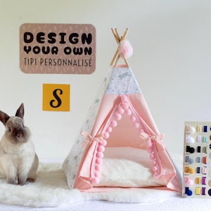 Pode incluir: Um pequeno tipi para animais com tecido rosa claro, acabamento em pompom e uma seção superior estampada. Um coelho castanho está sentado perto. Uma placa diz DESIGN YOUR OWN TIPI PERSONNALISÉ com um S abaixo. Há um mostruário de cores.