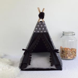 A black asanoha patterned teepee for rabbits and other small pets fitted with a gray faux sheepskin cushion next to a large jar filled with rabbit treats, dehydrated fruits and vegetables