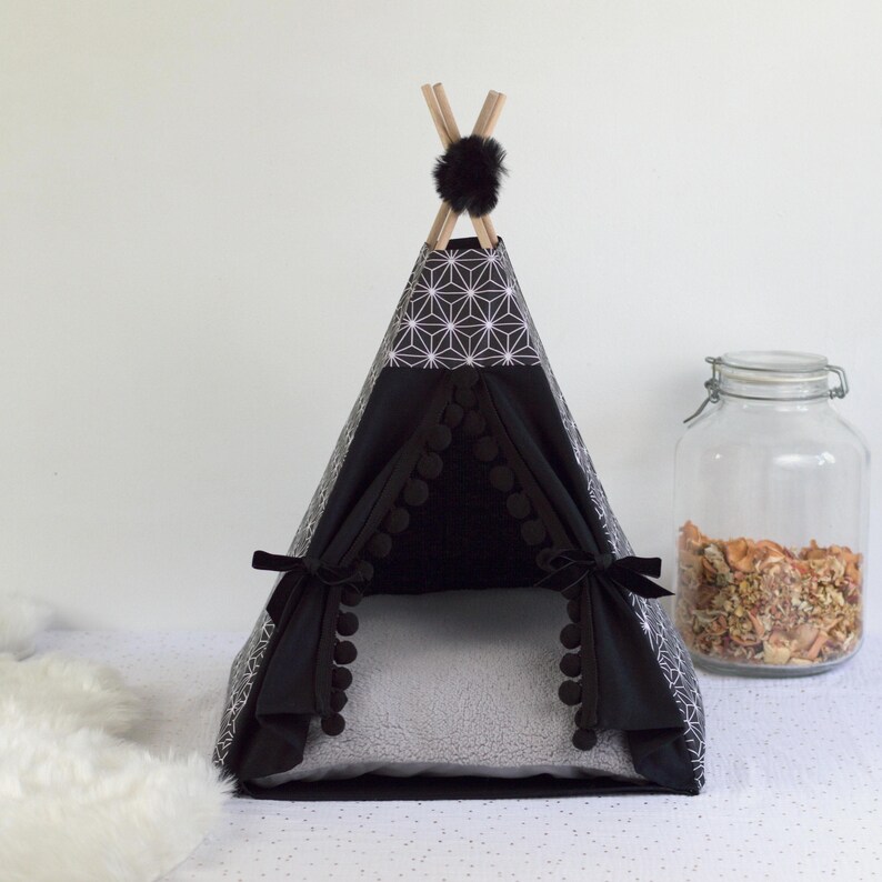 A black asanoha patterned teepee for rabbits and other small pets fitted with a gray faux sheepskin cushion next to a large jar filled with rabbit treats, dehydrated fruits and vegetables