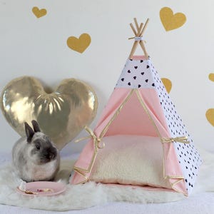 May include: A pink and white teepee-style pet bed with black heart accents, gold trim, and a fluffy interior. A gray rabbit sits on a white faux fur rug next to a pink plate. Gold heart decorations adorn the white background.