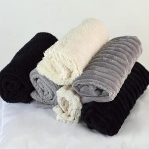 May include: Rolled plush blankets in black, grey, and cream are displayed. The blankets, with their soft textures and designs, are arranged on a white surface. The image showcases the cosy and inviting nature of the blankets, perfect for adding warmth and comfort.