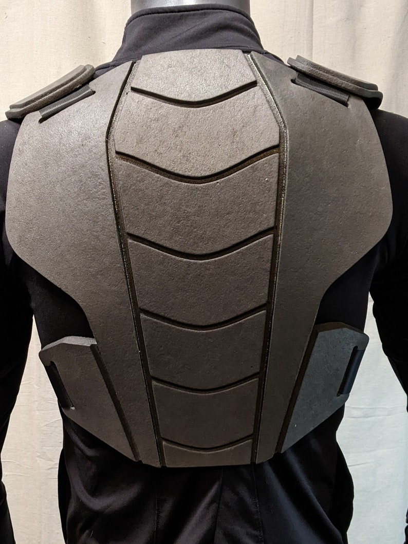 Puede incluir: Un protector de pecho acolchado negro con una superficie texturizada y una serie de paneles curvos en relieve. El protector tiene correas ajustables y est&aacute; dise&ntilde;ado para brindar protecci&oacute;n al pecho y la espalda.