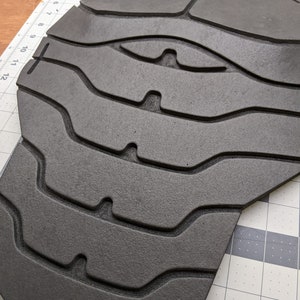 Puede incluir: Una pieza de armadura de espuma negra con una superficie texturizada y una serie de l&iacute;neas curvas elevadas. La pieza de armadura tiene forma de torso y tiene una parte superior redondeada.