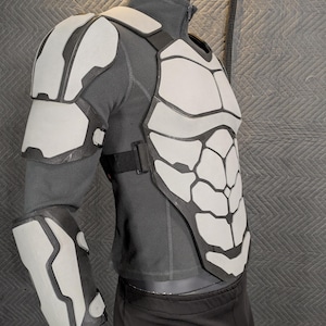 May include: A white and gray, futuristic-looking, padded armor chest piece with a matching shoulder pad and forearm guard. The armor is worn over a gray long-sleeved shirt and black pants.