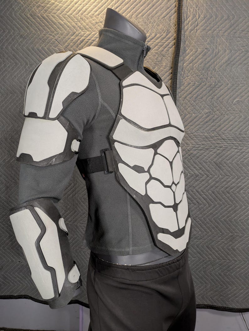 May include: A white and gray, futuristic-looking, padded armor chest piece with a matching shoulder pad and forearm guard. The armor is worn over a gray long-sleeved shirt and black pants.