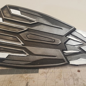 Pode incluir: Um escudo preto e prateado com um design geométrico. O escudo é feito de um material duro e tem uma superfície texturizada.