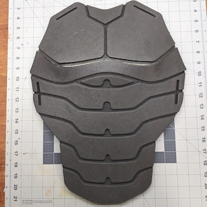 Puede incluir: Una pieza de espuma gris con un dise&ntilde;o texturizado, que se asemeja a un revestimiento de armadura. La pieza de espuma se coloca sobre una superficie blanca cuadriculada.
