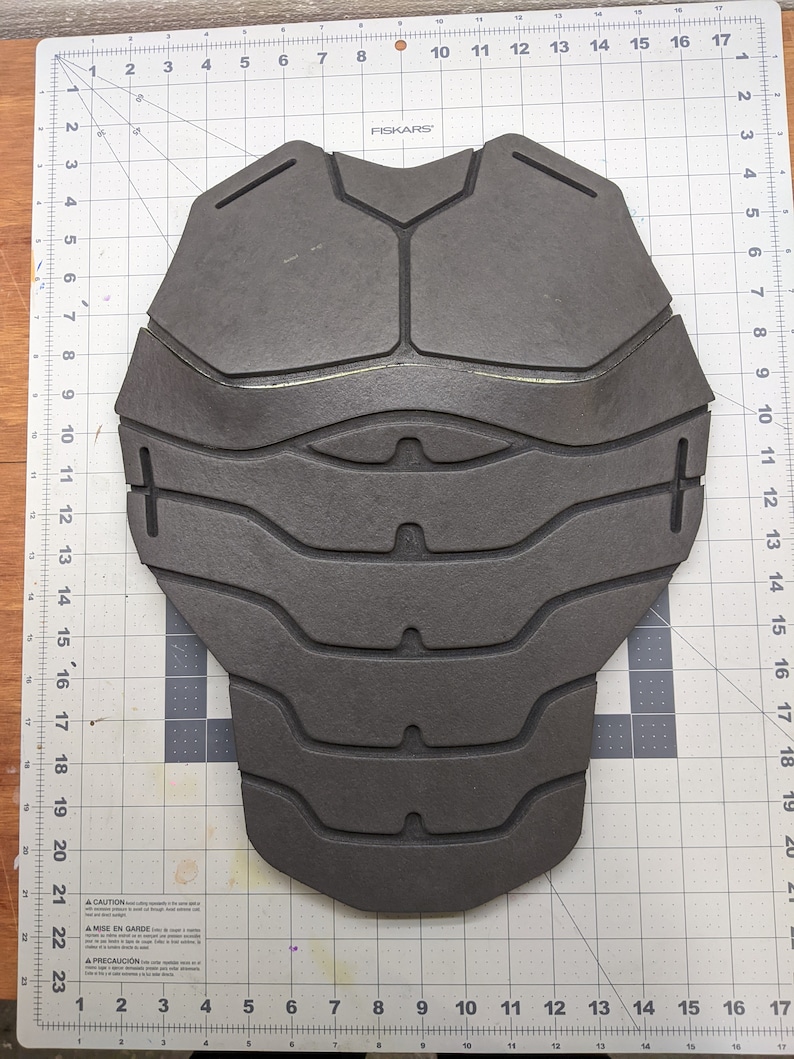 Puede incluir: Una pieza de espuma gris con un dise&ntilde;o texturizado, que se asemeja a un revestimiento de armadura. La pieza de espuma se coloca sobre una superficie blanca cuadriculada.