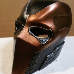 May include: A black and copper colored helmet with a detailed design. The helmet has a large eye opening and a smaller opening on the side. The helmet is made of a hard material and has a textured surface.