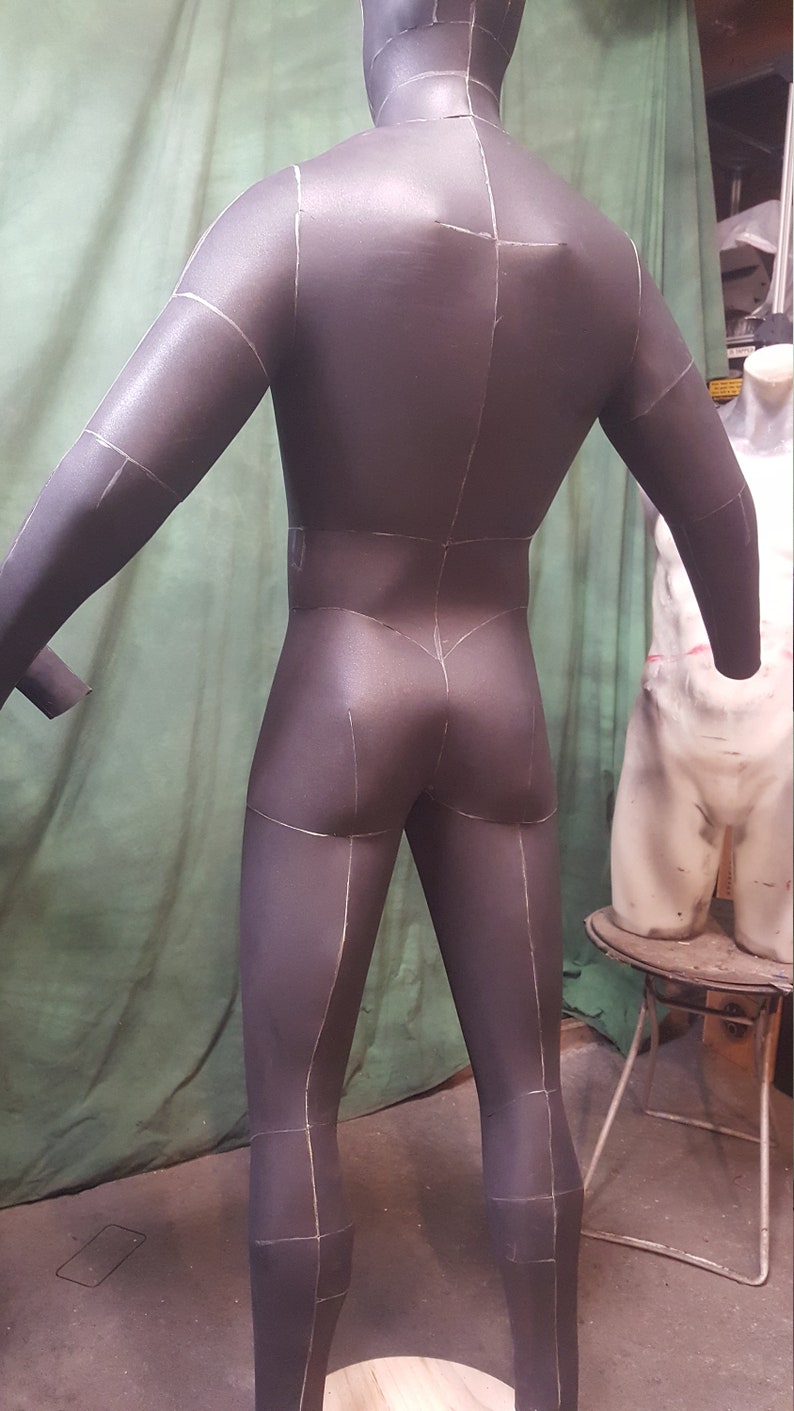 May include: A black foam mannequin with white lines drawn on it, showing the seams of a garment. The mannequin is standing on a wooden platform.