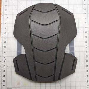 Puede incluir: Una pieza de armadura de espuma negra con una superficie texturizada y un dise&ntilde;o geom&eacute;trico. La pieza de armadura est&aacute; colocada sobre una superficie blanca cuadriculada.