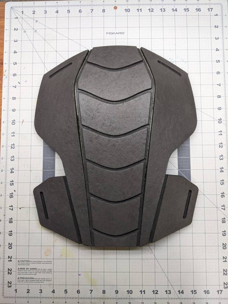 Puede incluir: Una pieza de armadura de espuma negra con una superficie texturizada y un dise&ntilde;o geom&eacute;trico. La pieza de armadura est&aacute; colocada sobre una superficie blanca cuadriculada.