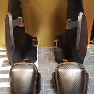 May include: A pair of black knee pads with a gray and black checkered pattern and a copper-colored trim. The knee pads are on a gray cutting mat.